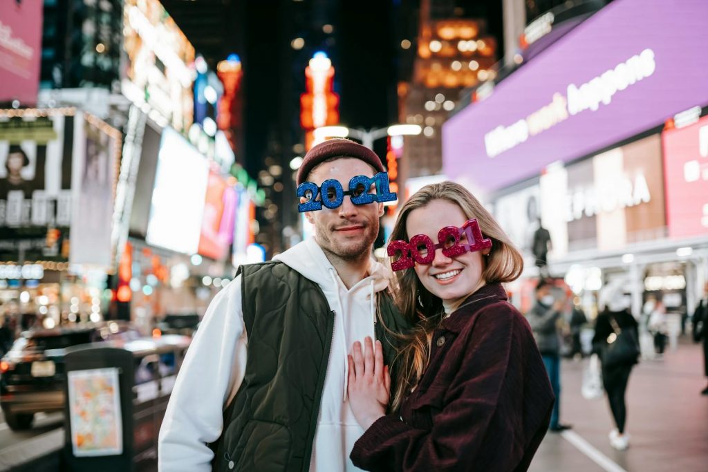 Cheerful couple wearing decorated 2021 glasses looking at camera and embracing while standing on square with glowing modern buildings on blurred background at night time