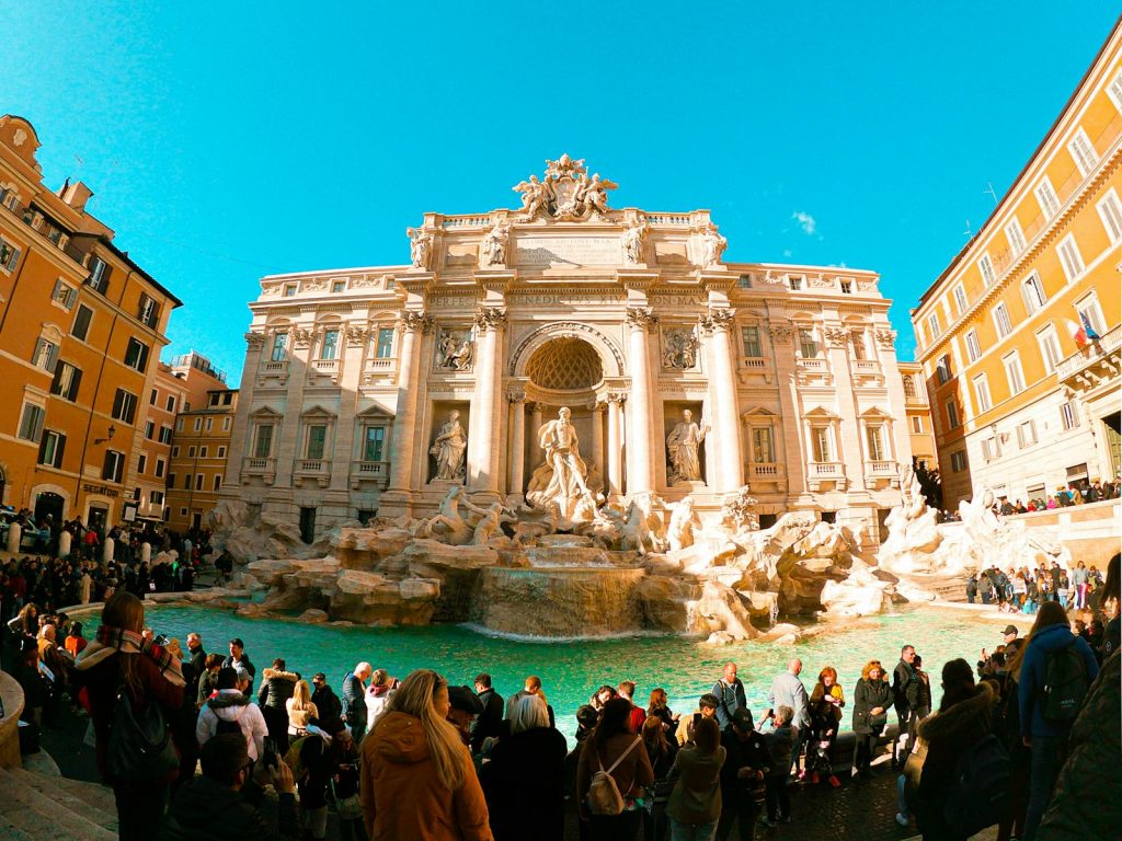 A large crowd gathers at the historic Trevi Fountain in Rome, Italy, basking in the sunlight.
