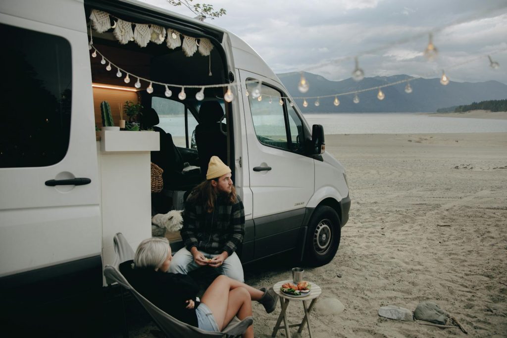 Couple enjoying beachside relaxation next to camper van with string lights under cloudy skies.