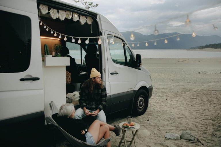 Couple enjoying beachside relaxation next to camper van with string lights under cloudy skies.