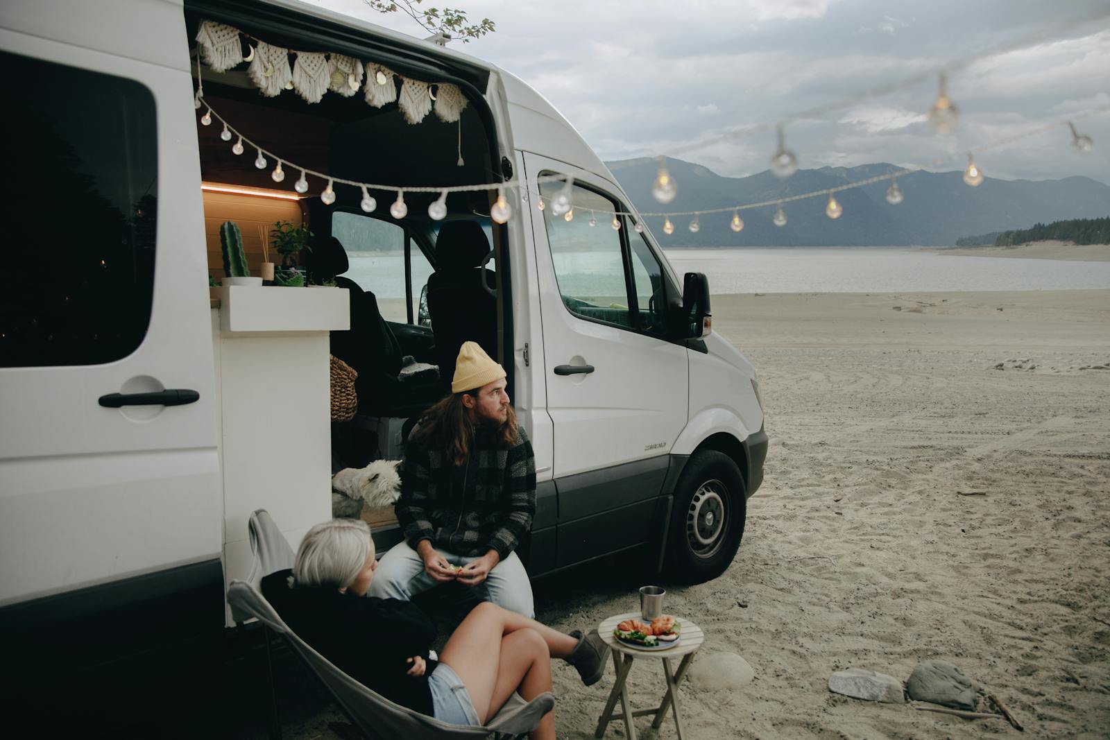 Couple enjoying beachside relaxation next to camper van with string lights under cloudy skies.