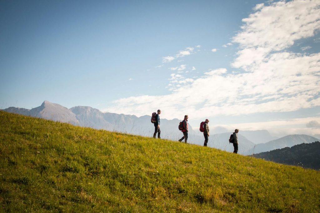 Four hikers with backpacks walking downhill on a sunny day with mountains in the background.