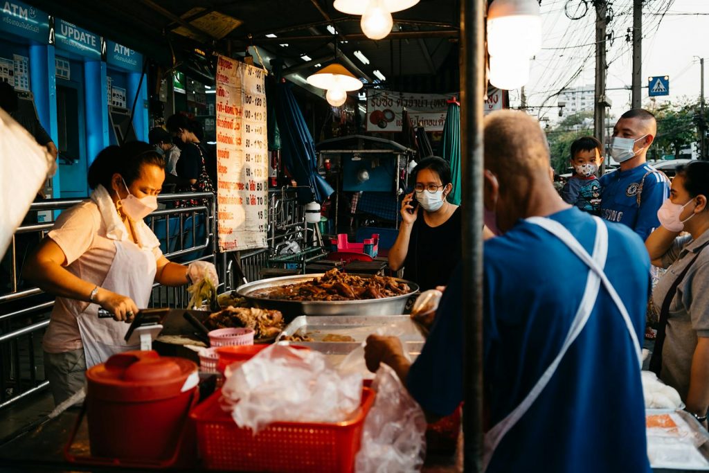 Market vendor selling food at a street market in Bangkok, with people wearing face masks.
