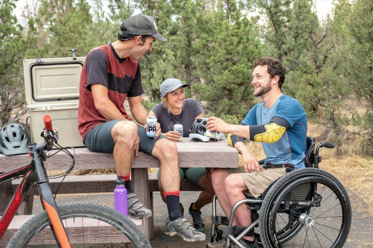 Group of friends having fun outdoors, chatting and enjoying drinks by a picnic table.