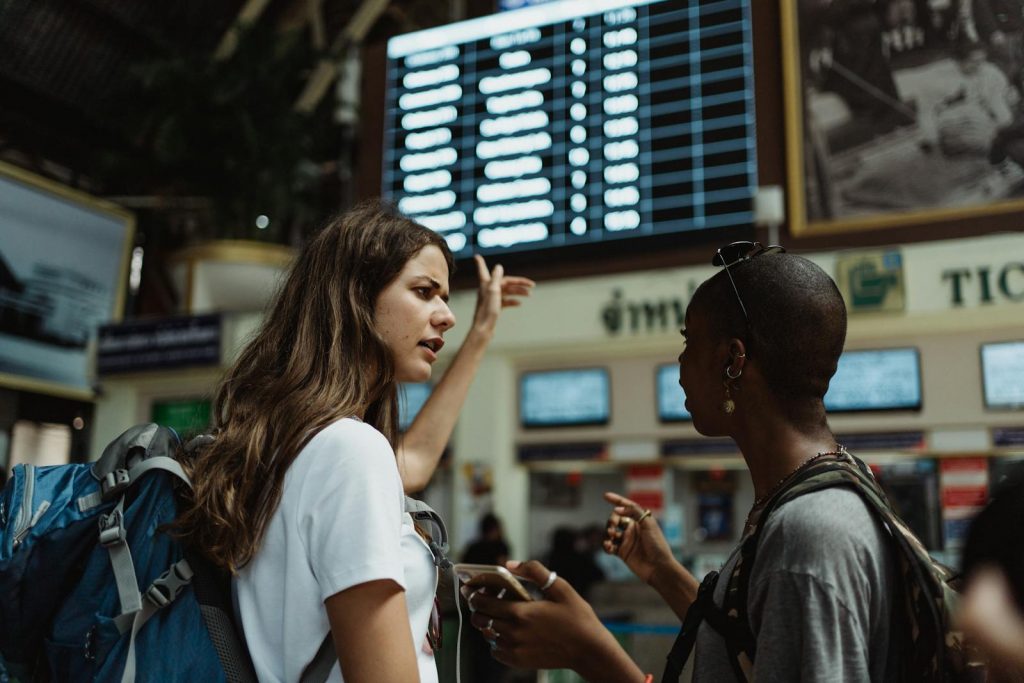 Two travelers with backpacks discuss in front of an airport timetable. Adventure awaits.