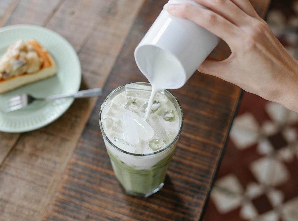 Close-up of a creamy iced matcha latte being poured, accompanied by a delicious slice of cake on a wooden table.