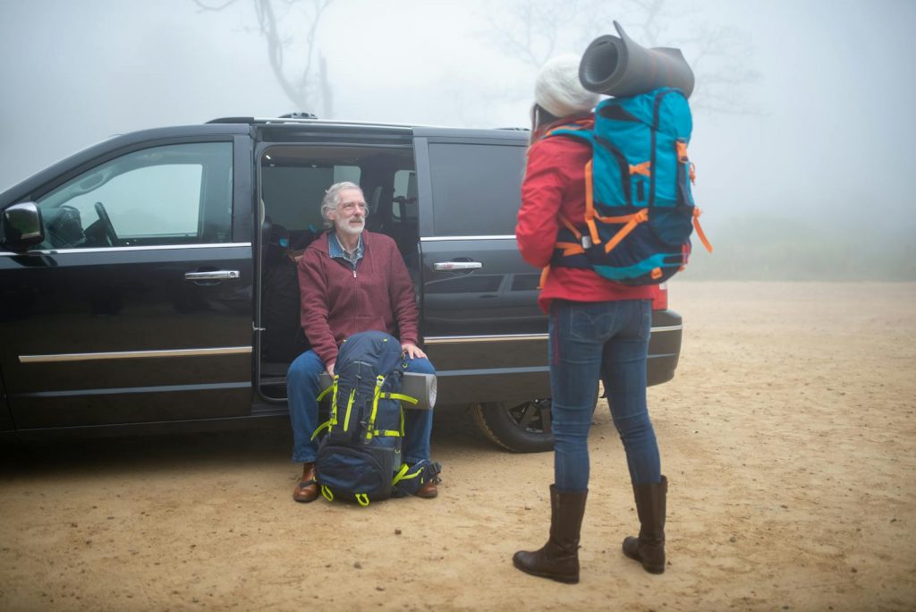 An elderly couple with backpacks getting ready for a foggy hiking trip near a vehicle.