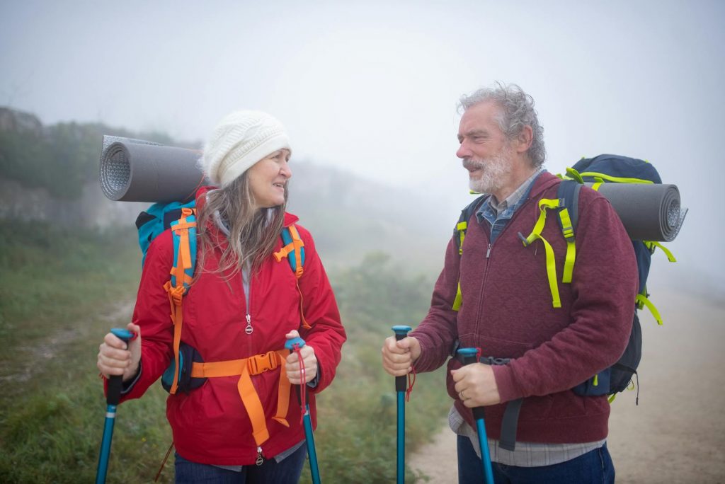 A senior couple hiking with trekking poles amidst foggy mountain landscape in Portugal, sharing a joyful moment.
