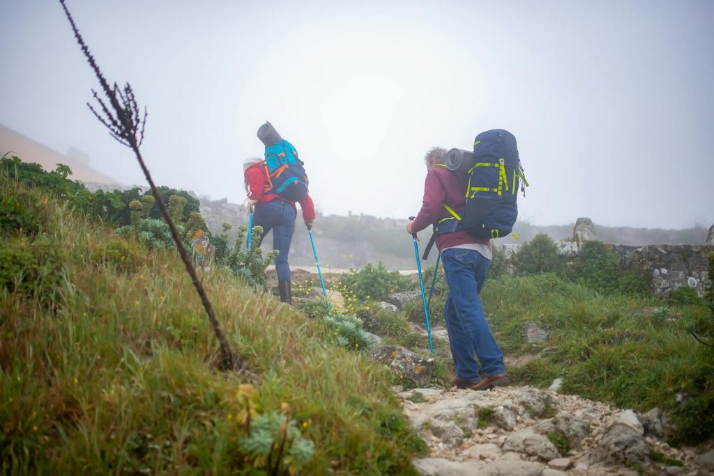 A senior couple enjoys a misty hike in Portugal, each carrying backpacks and walking sticks.
