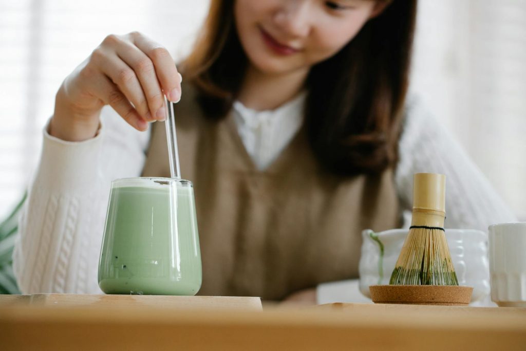 A woman stirs an iced matcha latte with a straw, using a traditional matcha whisk.