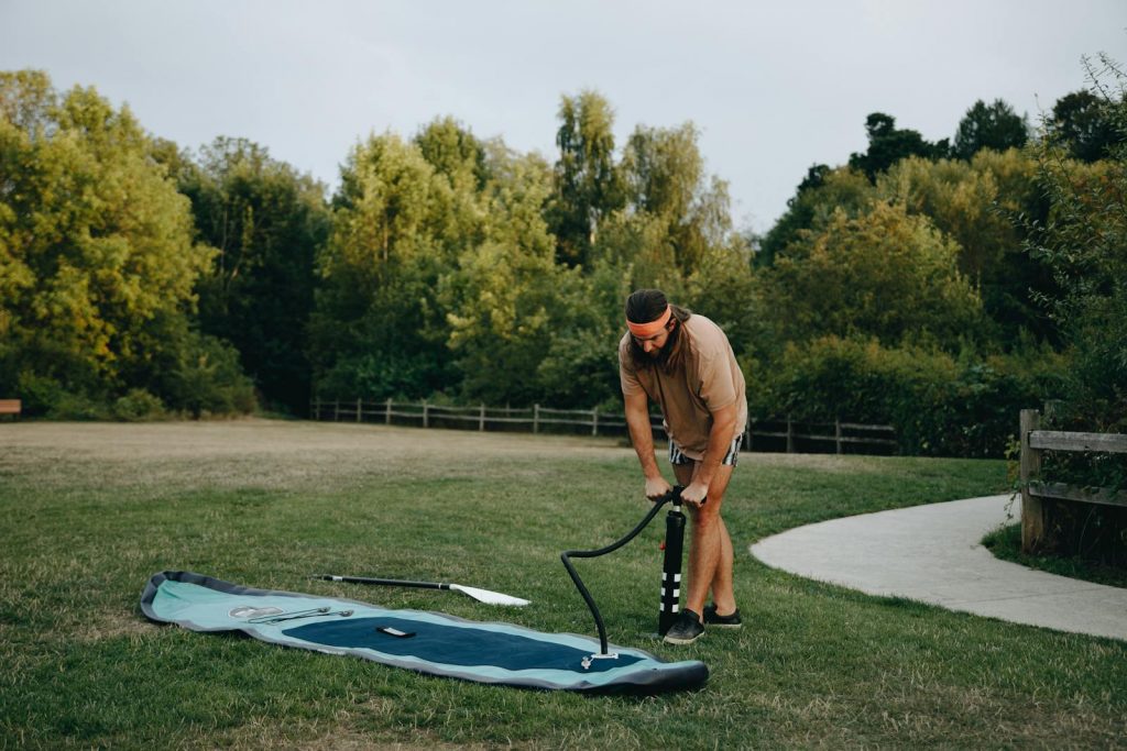 Man using pump to inflate paddle board in a sunny park, surrounded by trees.
