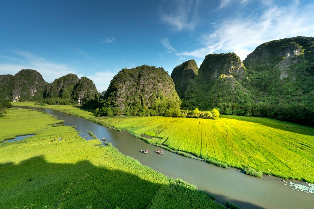 Aerial view of a scenic river cutting through lush green fields with towering limestone mountains under a clear sky.