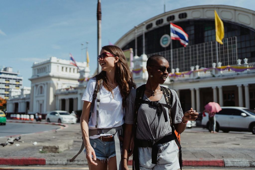 Two women smiling and walking outside a railway station in Bangkok on a sunny day.