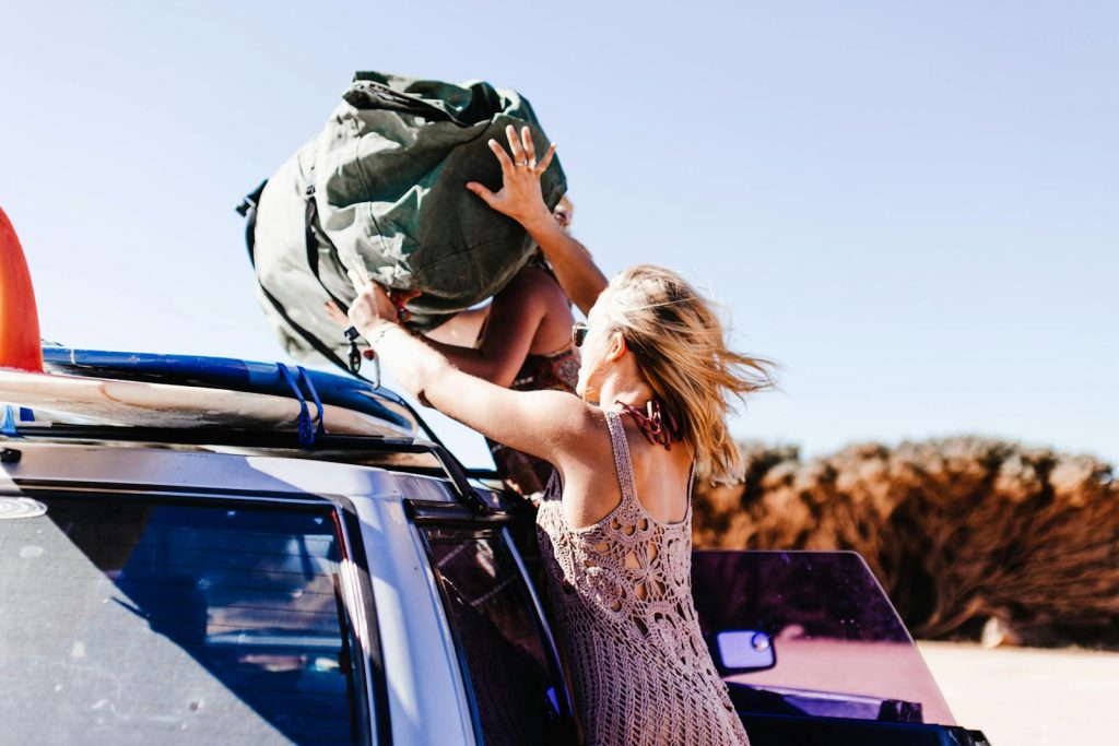 A woman placing a large bag on a car's roof rack, symbolizing summer travel adventure.