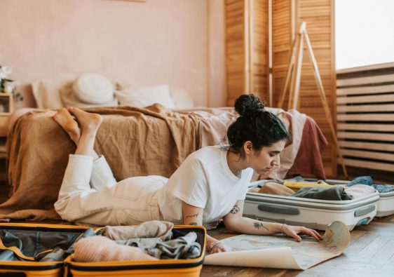 Woman lying on floor planning a vacation with a map and suitcases in a cozy bedroom.