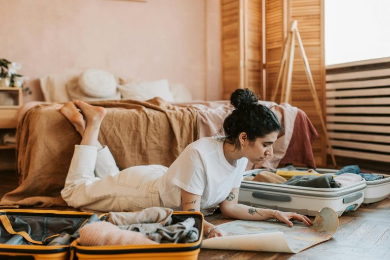 Woman lying on floor planning a vacation with a map and suitcases in a cozy bedroom.