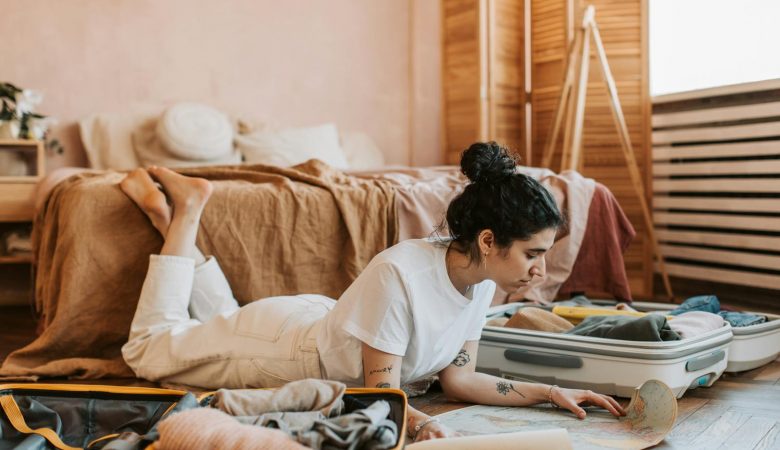Woman lying on floor planning a vacation with a map and suitcases in a cozy bedroom.