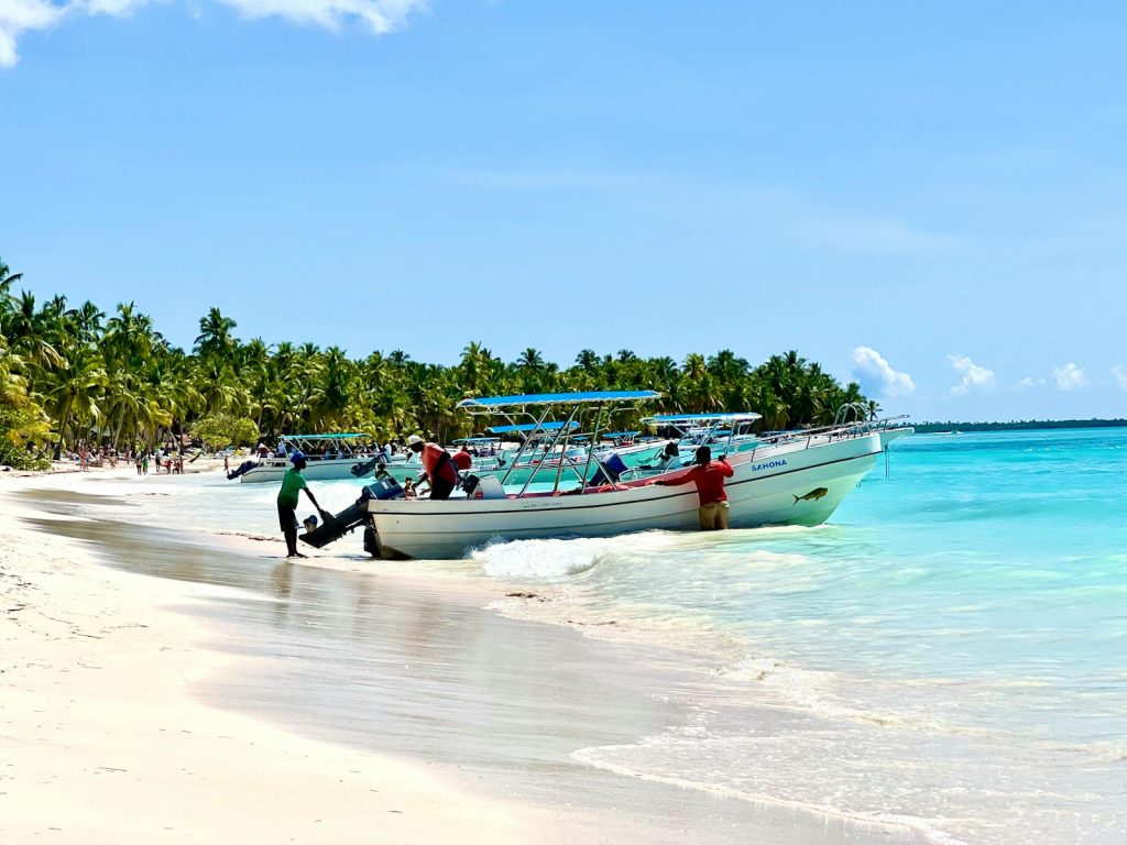 Vibrant scene of boats docked on a sunny beach in the Dominican Republic, surrounded by clear turquoise water and palm trees.