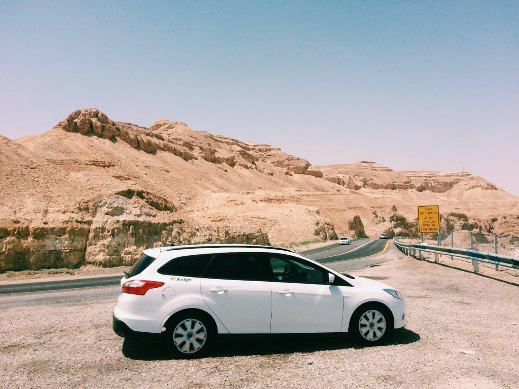 White car parked by a desert highway with scenic desert mountains and clear blue sky in the background.
