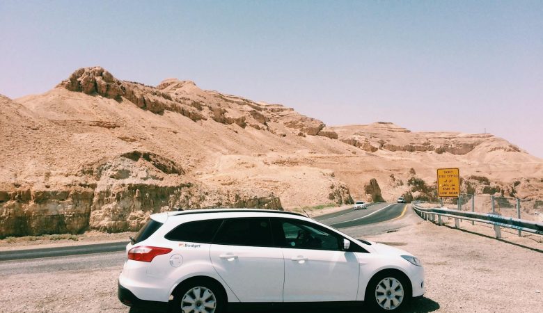 White car parked by a desert highway with scenic desert mountains and clear blue sky in the background.