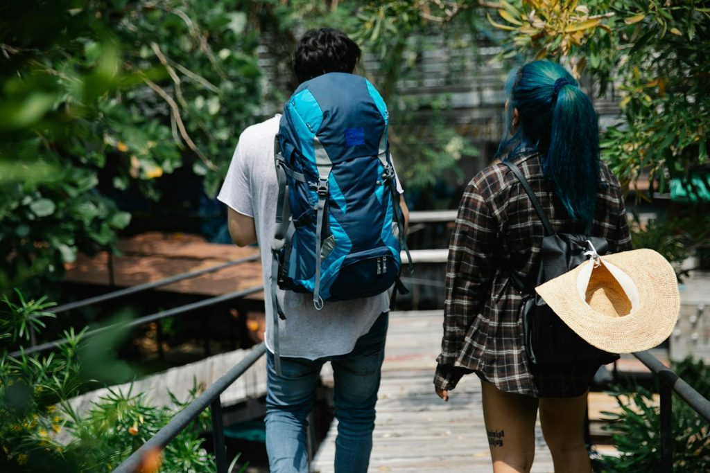 Young couple walks through forest with backpacks and hats, enjoying sunlight and nature.
