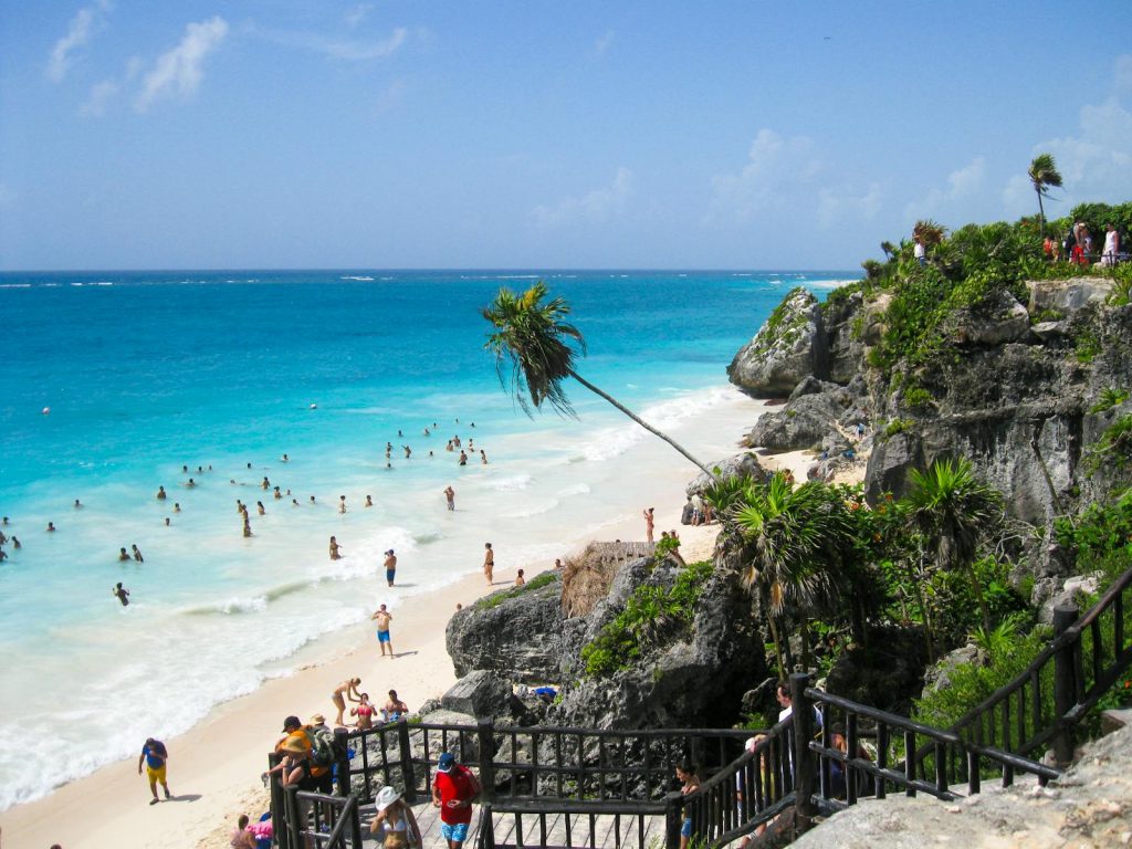 Crowded beach with turquoise water and rocky cliffs under a bright sky.