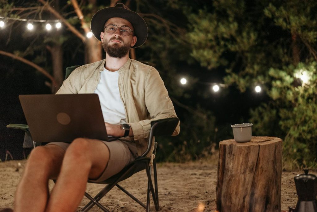 Bearded man enjoying a relaxing night outdoors using a laptop with string lights in the background.