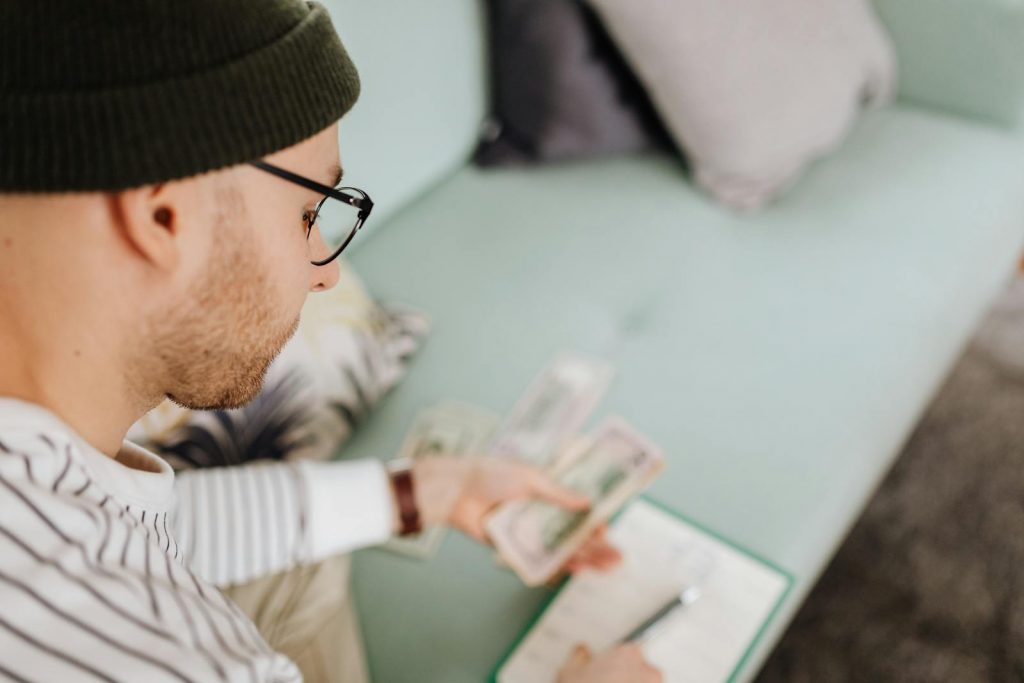 Adult man wearing glasses and beanie counting cash while budgeting indoors.