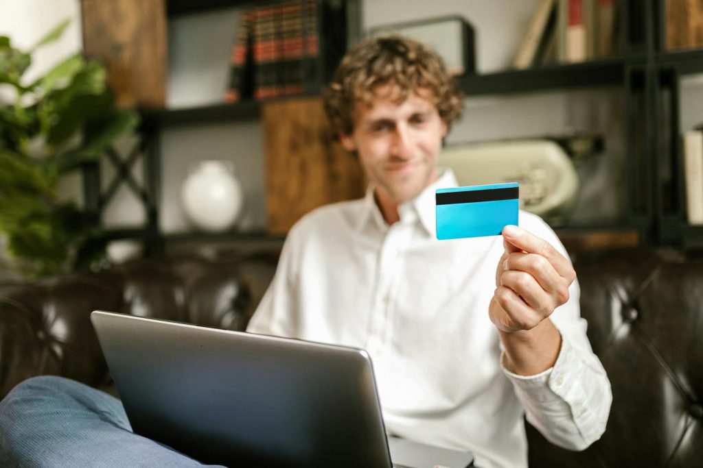 Smiling man with curly hair sits on couch holding a credit card for online shopping.