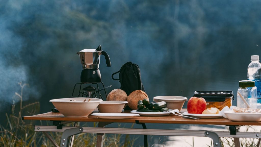 Enjoy a tranquil outdoor breakfast by the lake with a classic coffee maker and fresh ingredients on a camping table.
