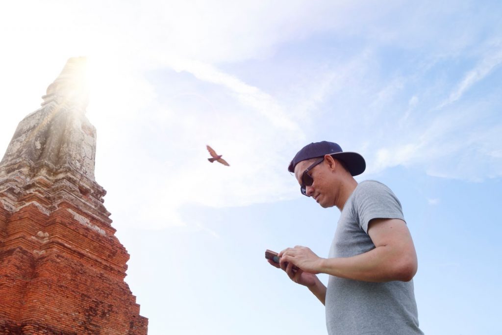 Ein Mann mit Sonnenbrille schaut in der Nähe eines antiken Tempels auf sein Handy; der Himmel ist klar und blau, und ein Vogel fliegt darüber.