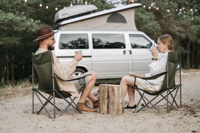 A couple relaxes with coffee outside their van, surrounded by nature, enjoying a camping trip.